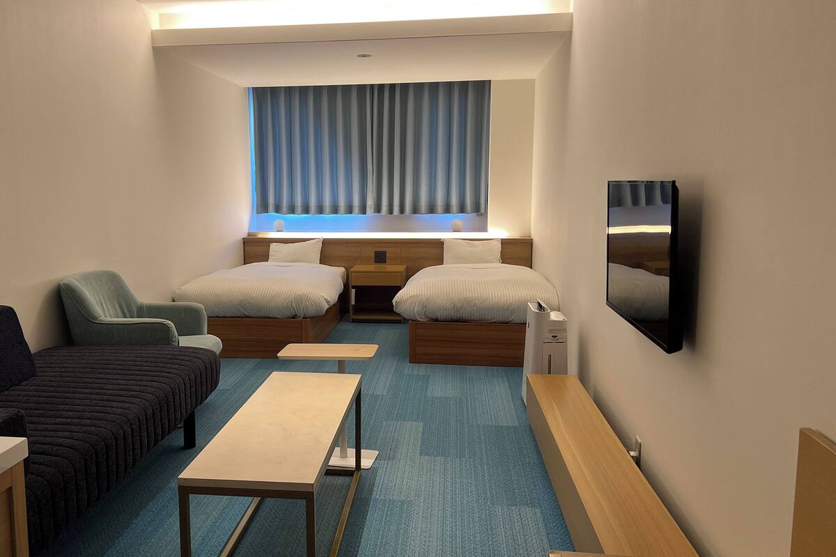 A hotel room with two beds, a sofa, a small table, a TV, and a window with curtains.