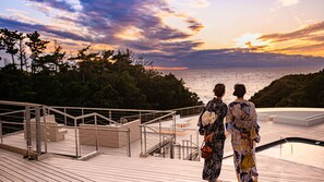 Twee personen staan op een houten terras met uitzicht op een wateroppervlak, met een zonsondergang op de achtergrond.