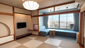 Quarto com piso de tatame, uma mesa baixa, um sofá, uma TV e janelas amplas com cortinas.