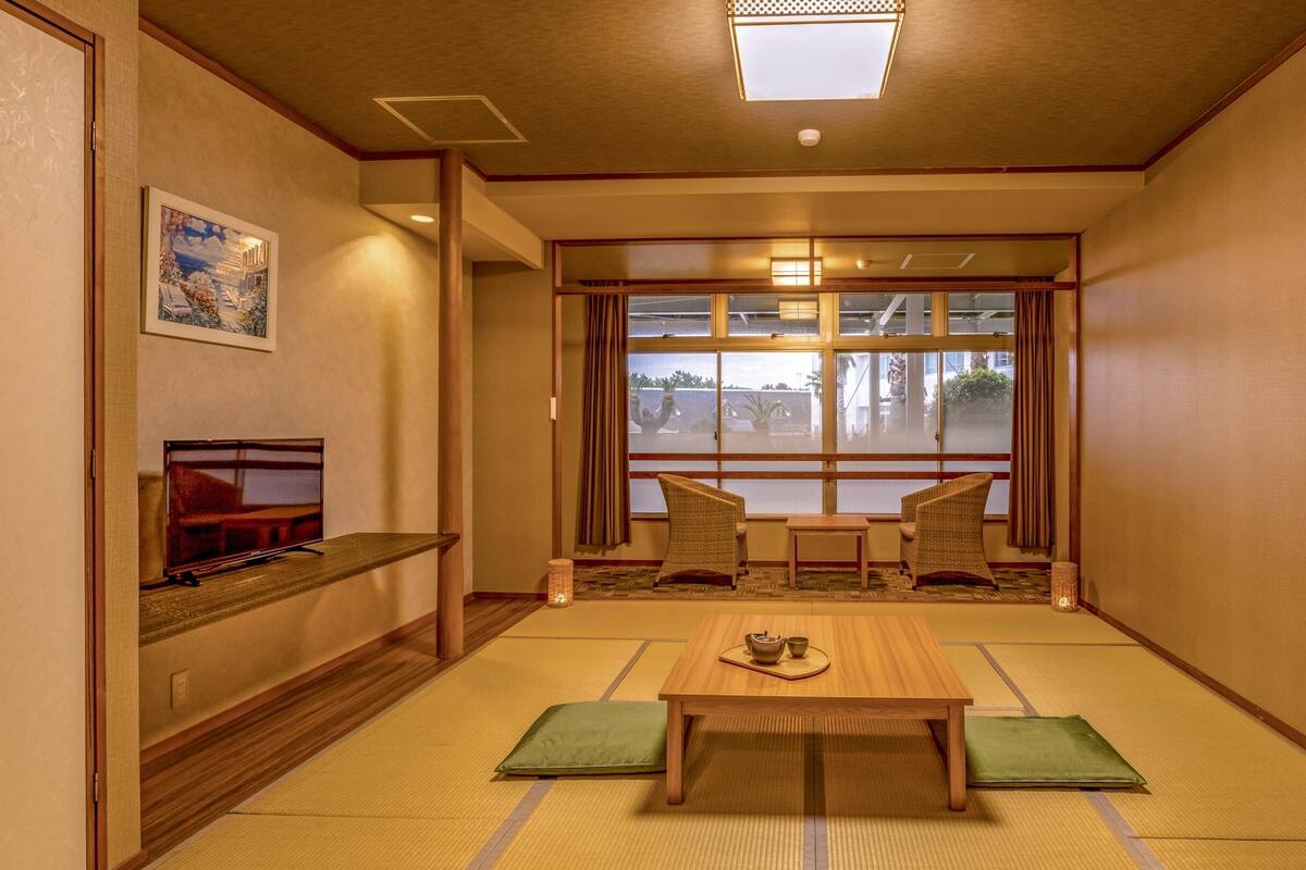 A traditional Japanese-style room with tatami flooring, a low table, two chairs, and a large window with a view of the outside.