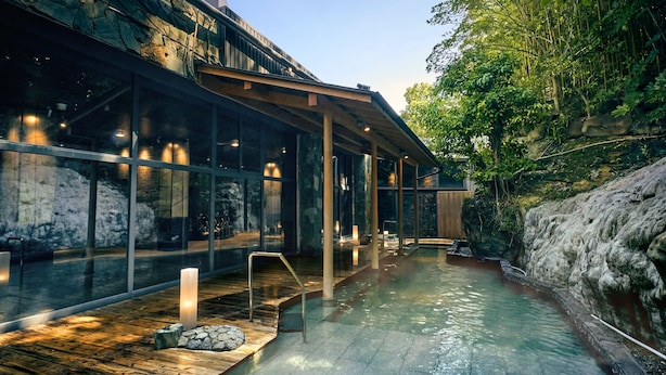 Un área de spa con terraza de madera, un manantial termal y una pared de vidrio que refleja la naturaleza circundante.