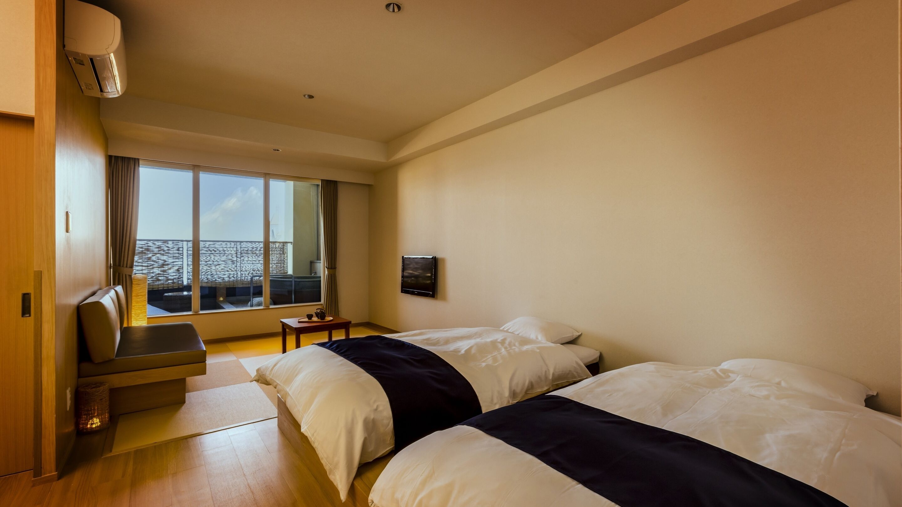 A hotel room with two beds, a small table, a chair, a window with a view, and an air conditioning unit.
