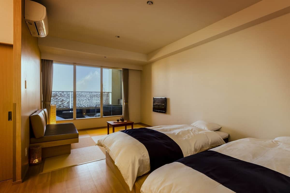 Een hotelkamer met twee bedden, een tafeltje, een stoel, een raam met uitzicht op zee, en een airconditioning.