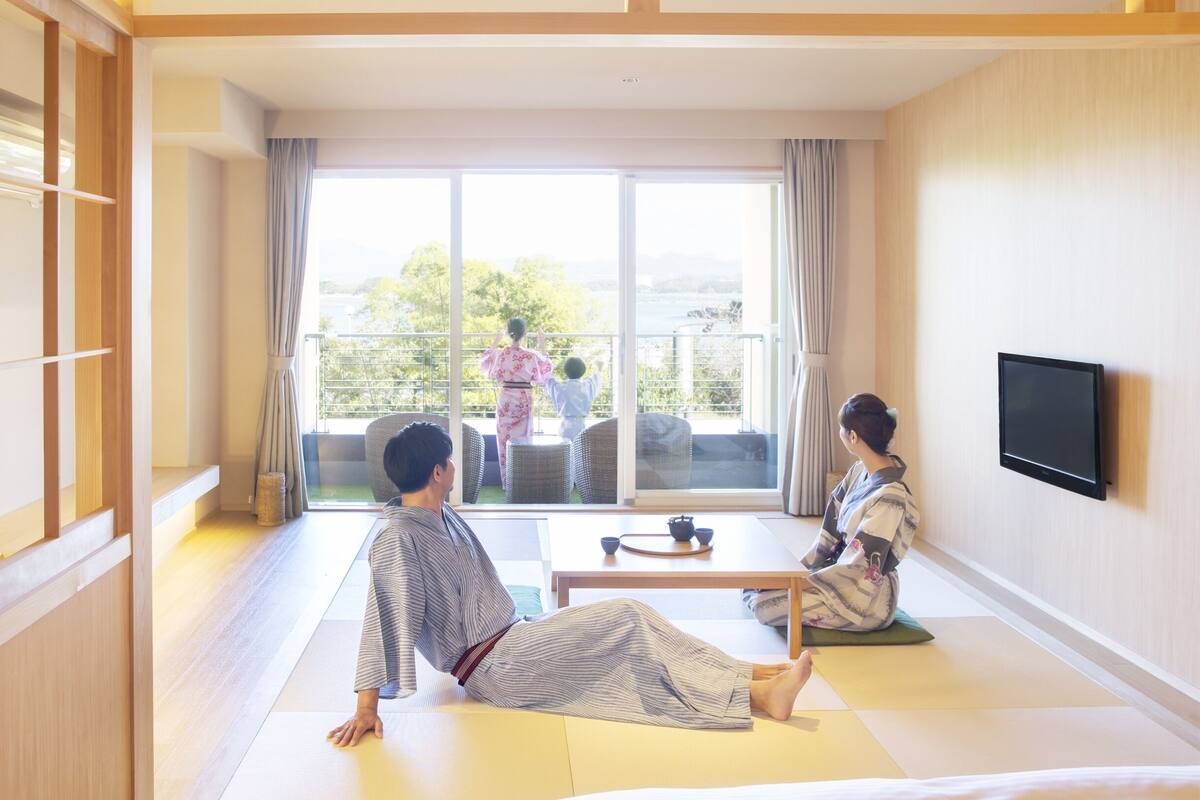 Een traditionele Japanse kamer met tatami vloerbedekking, een schuifdeur naar een balkon en een flatscreen televisie aan de wand.