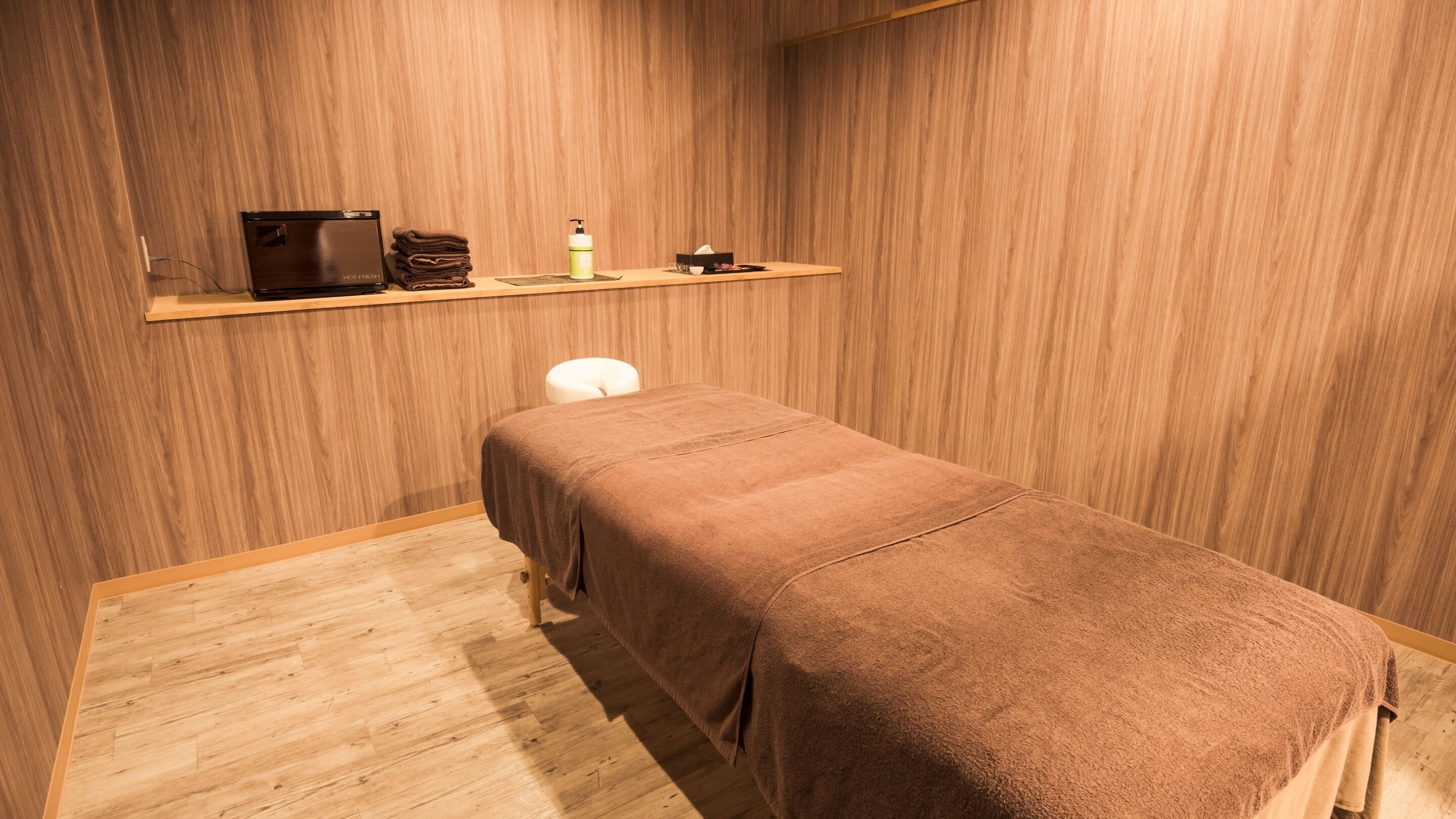 Una habitación con piso de madera, una cama individual con manta marrón, un estante pequeño con artículos de tocador y una silla blanca.