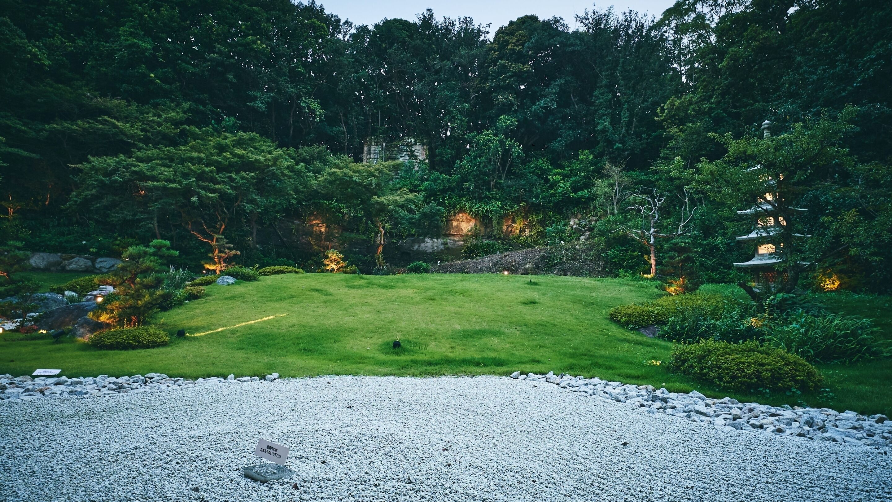 A serene garden with a gravel path, lush greenery, and traditional structures.