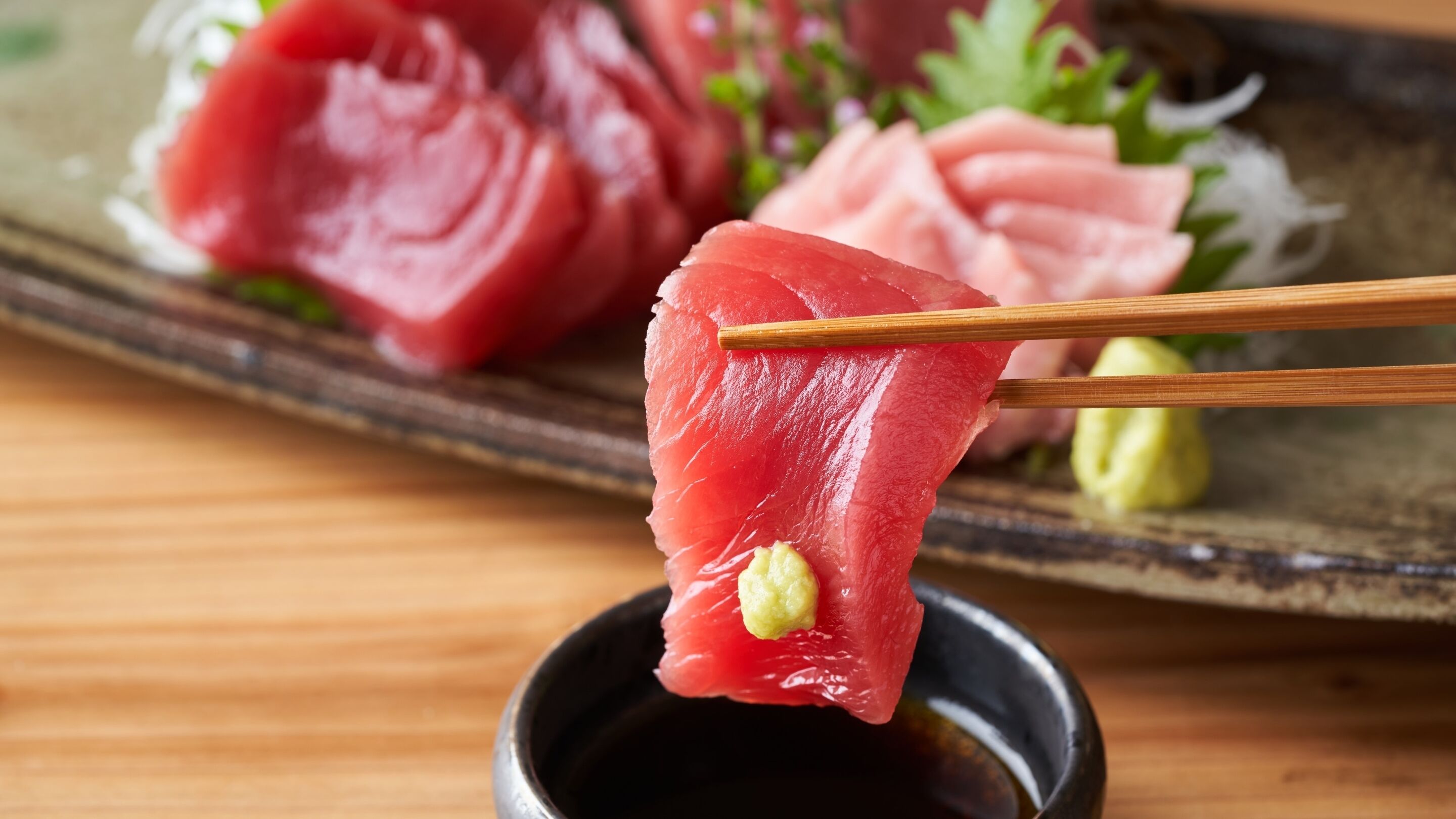 Une assiette de sashimi, avec des baguettes tenant un morceau, et un petit bol de sauce soja.