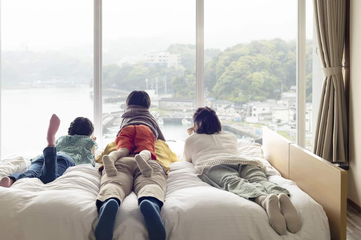 Drei Personen liegen auf einem Bett und blicken durch ein großes Fenster auf ein Gewässer und Gebäude.