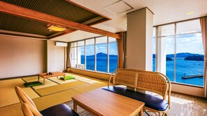 Une pièce avec des tatamis, une table en bois, une chaise avec un coussin et de grandes fenêtres offrant une vue sur la mer et les montagnes.