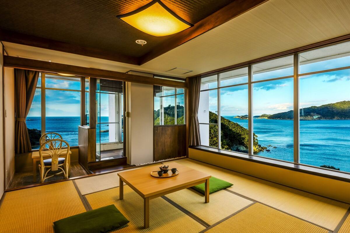 Ein Zimmer mit Tatami-Böden, einem niedrigen Tisch und Blick auf das Meer und die Hügel.