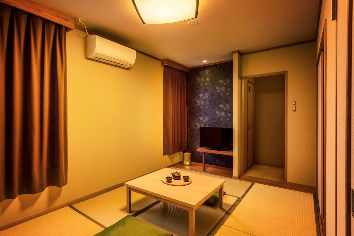 Ein traditioneller japanischer Raum mit Tatami-Böden, einem niedrigen Tisch mit Teeservice, einem Fernseher auf einem Standfuß und einer Schiebetür.