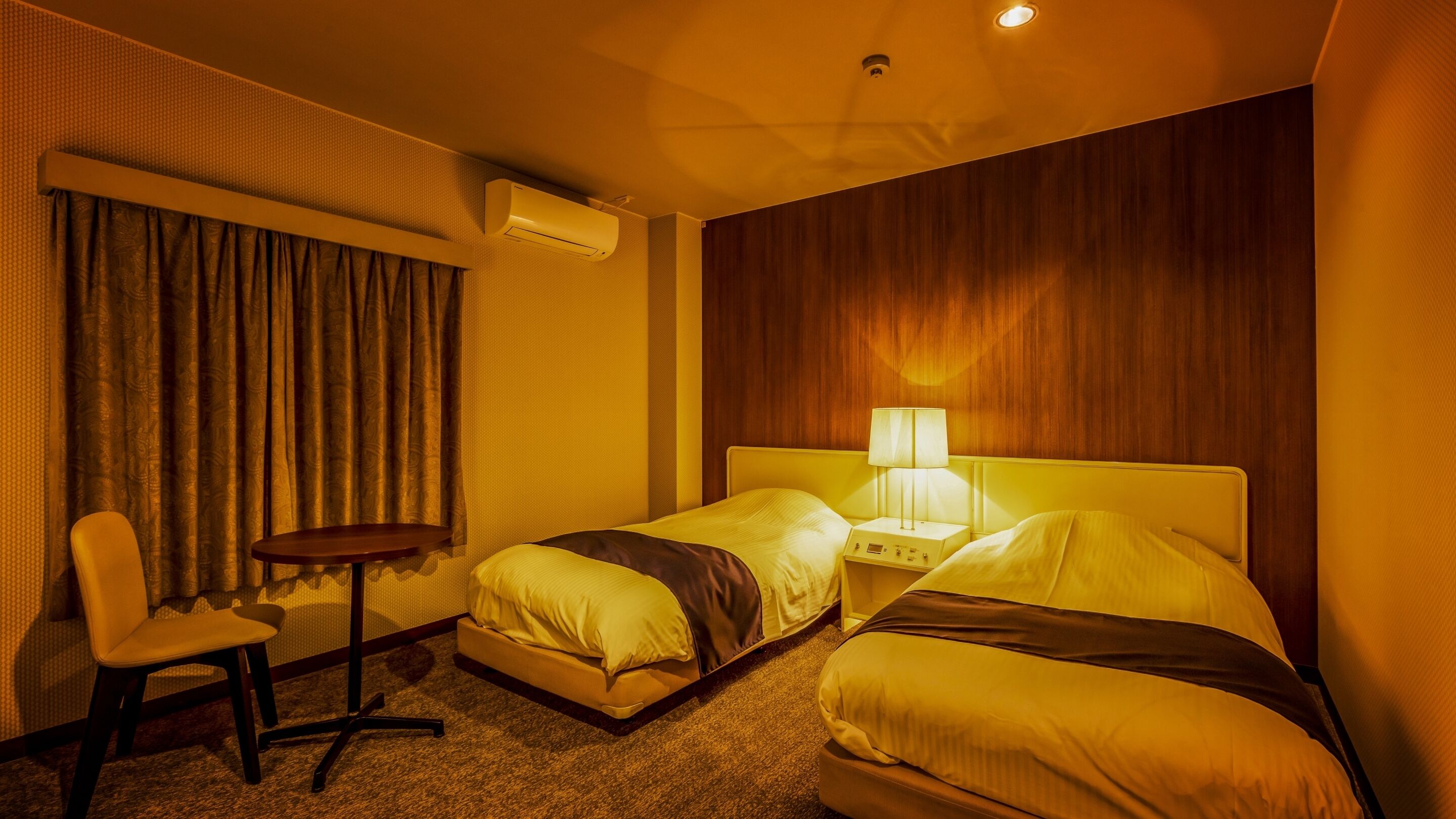 Quarto de hotel com duas camas, uma escrivaninha, uma cadeira, um aparelho de ar condicionado e um abajur.