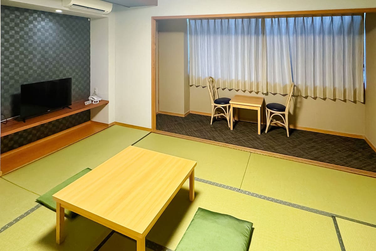 Ein Zimmer mit Tatami-Matten, einem Fernseher, einem Tisch und einem Vorhangfenster.