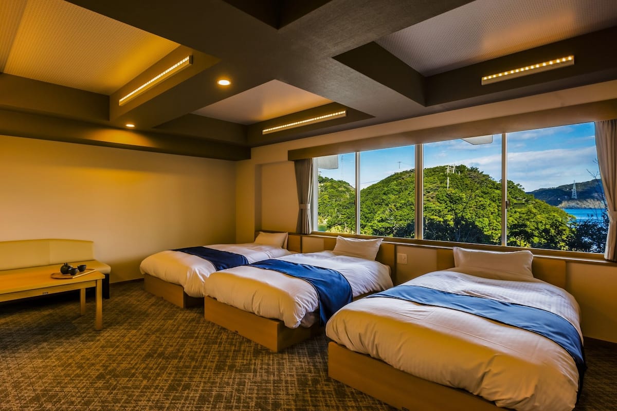 Ein Hotelzimmer mit zwei Betten, einer Bank und einem großen Fenster mit Blick auf eine grüne Landschaft.
