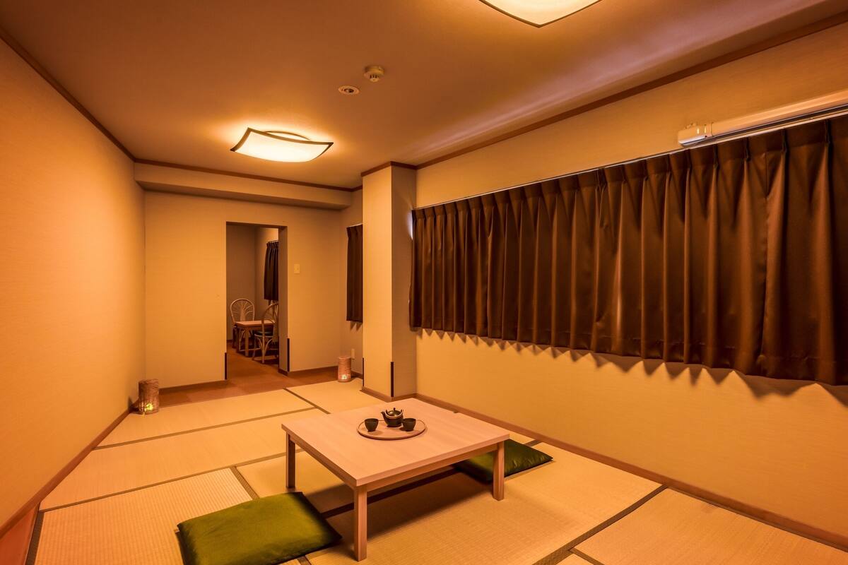 Ein Zimmer mit Tatami-Boden, einem niedrigen Tisch mit Tablett, einem Fenster mit dunklen Vorhängen und einem Gang zu einem anderen Zimmer.