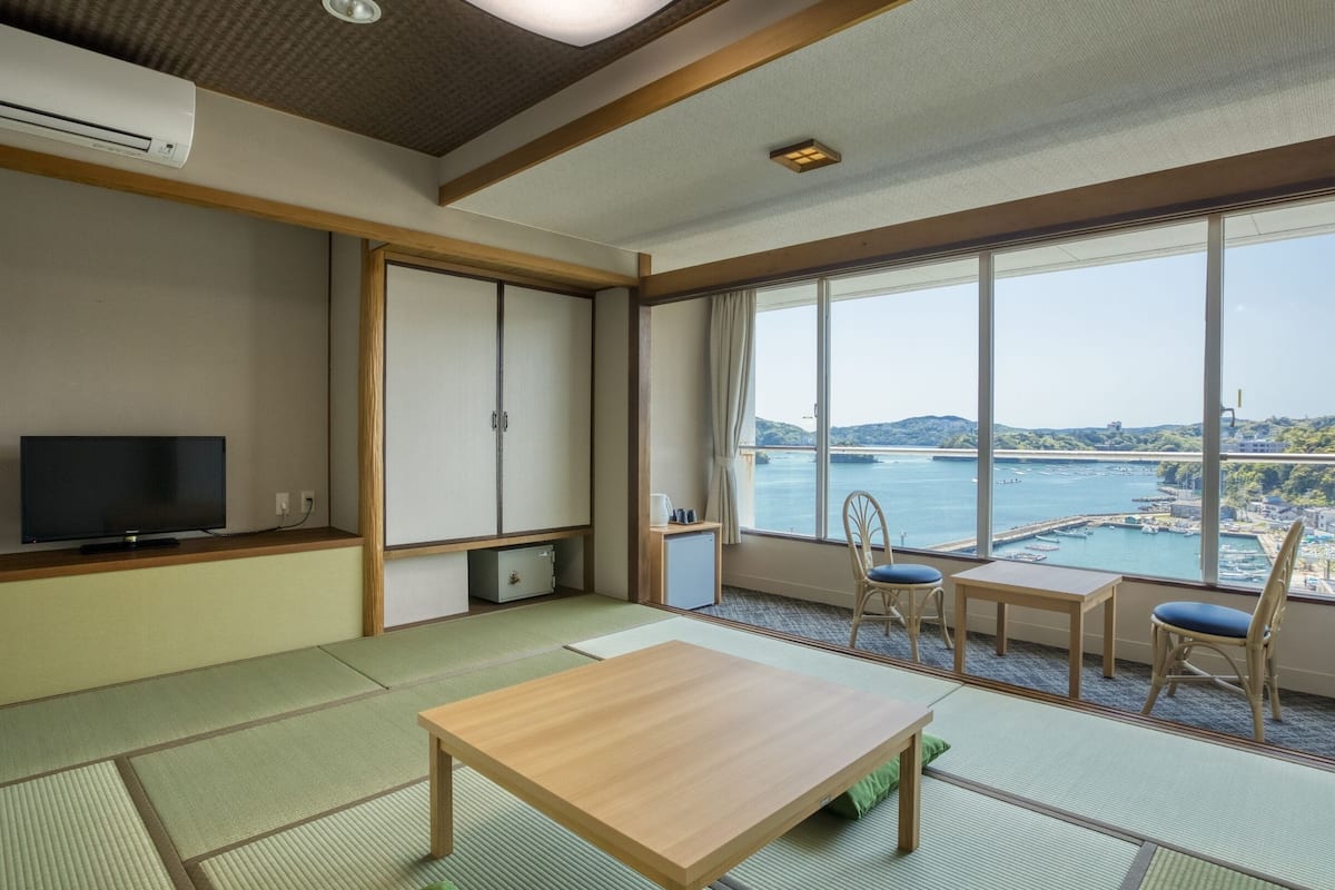 Ein Zimmer mit Tatami-Matten, einem Fernseher, einem Tisch, Stühlen und einem großen Fenster mit Blick auf ein Gewässer und eine Brücke.