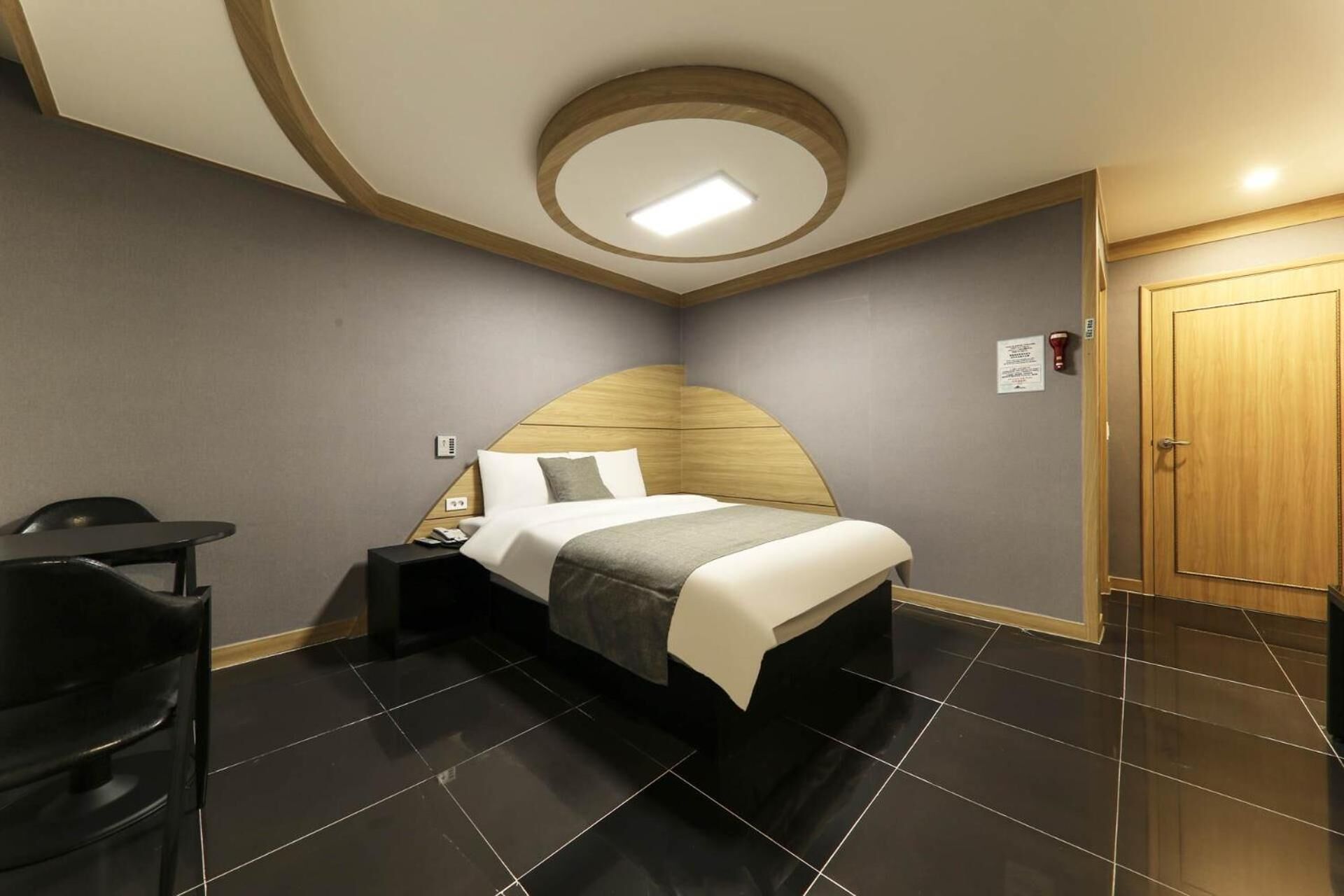 A modern hotel room with a curved ceiling, a bed with a wooden headboard, a desk, and a door.