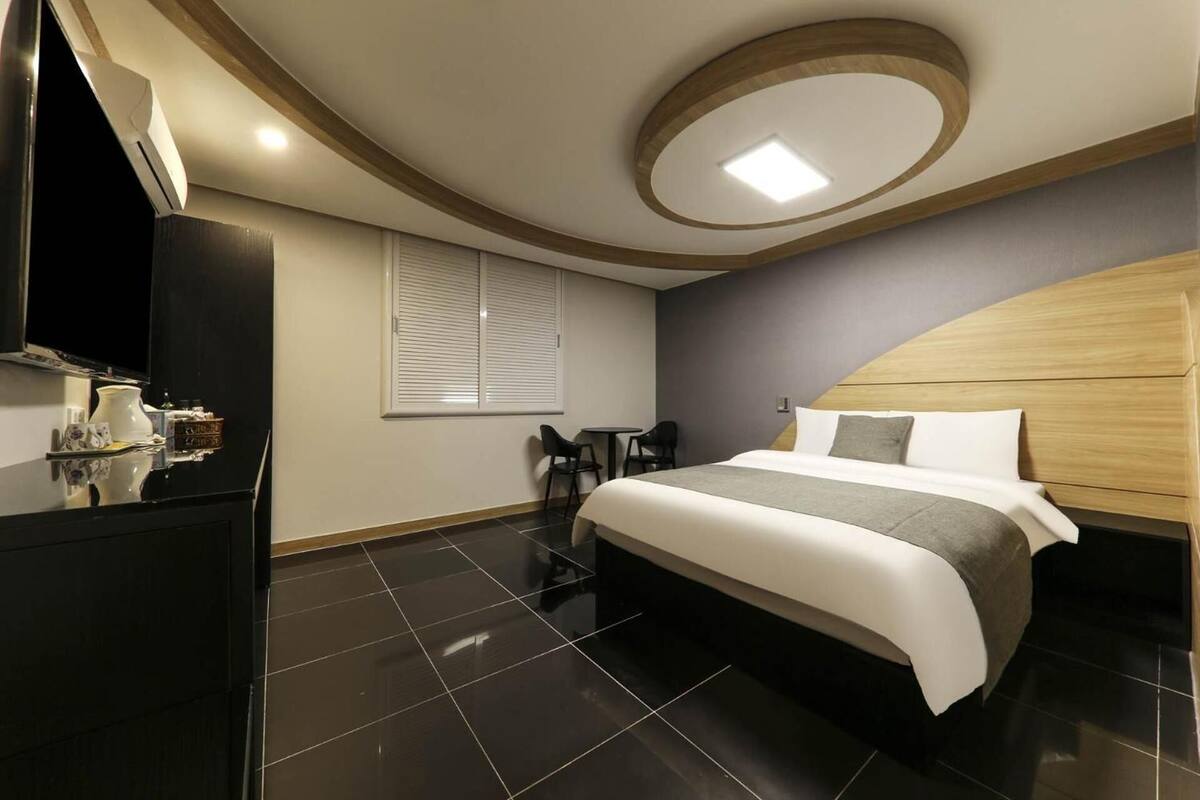 A modern hotel room with a large bed, a TV, a small table, and a window with blinds.