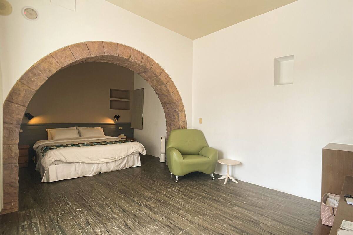 Ein Schlafzimmer mit einem Steingewölbe, einem Bett mit weißer Bettwäsche, einem grünen Sessel, einem kleinen runden Tisch und einer hölzernen Kommode.