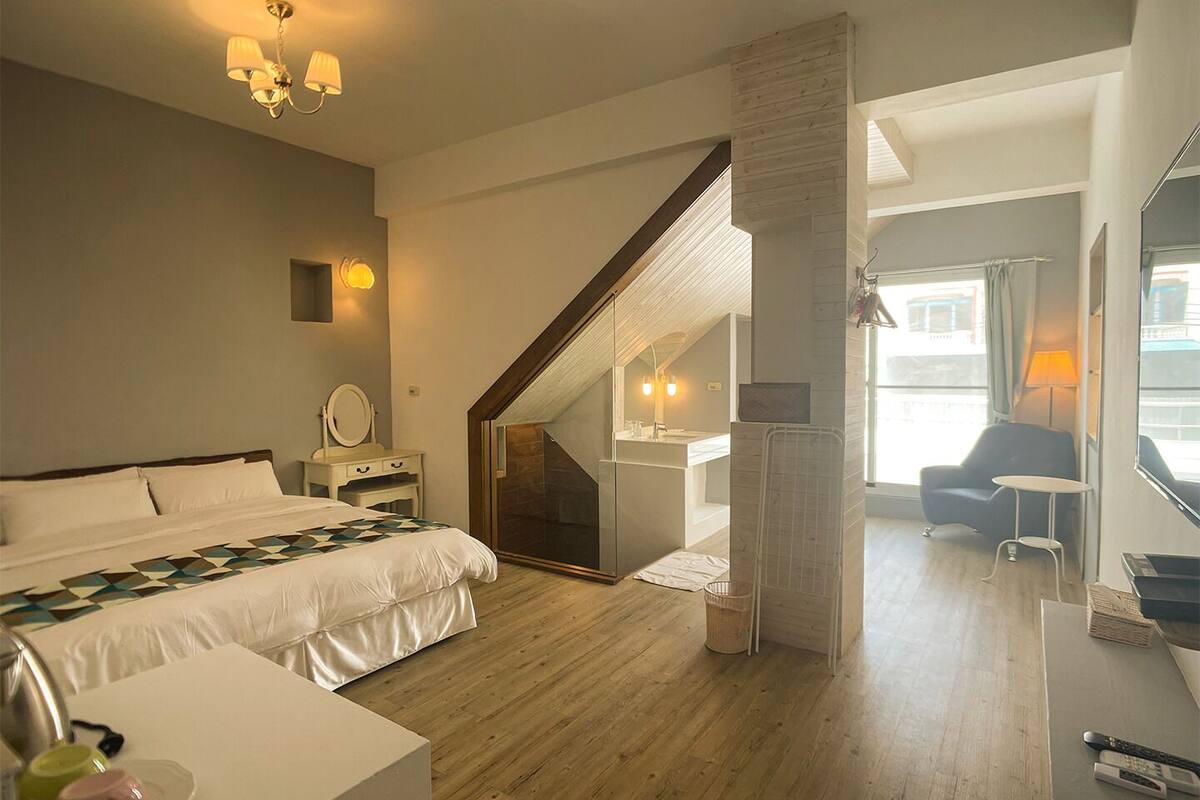 Ein Hotelzimmer mit Bett, Nachttisch, Lampe und Blick auf die Stadt durch große Fenster.