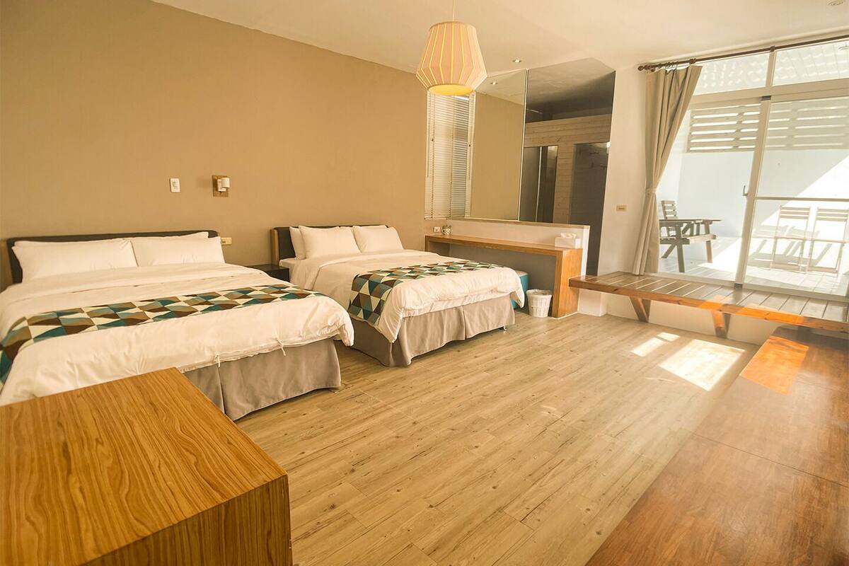 Ein Hotelzimmer mit zwei Betten, einer Holzbank, einem Schreibtisch und einem großen Fenster mit Blick ins Freie.