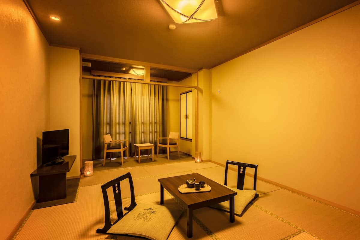 Ein kleiner, gemütlicher Raum mit Tatami-Boden, einem niedrigen Tisch mit Teeservice, zwei Stühlen, einem Fernseher und einer Schiebetür, die auf einen Balkon führt.