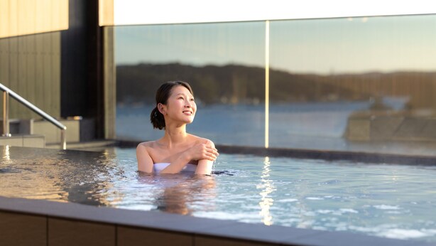 A person in a hot tub with a view of a body of water and a building in the background. - Ooedo Onsen Monogatari Premium Shima Saichoraku (Shima)