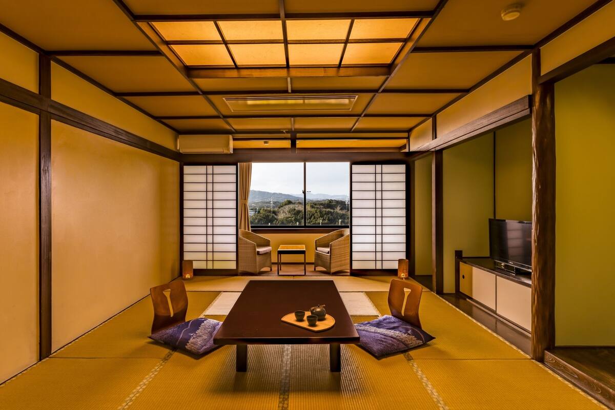 Ein traditioneller japanischer Raum mit Tatami-Bodenbelag, einem niedrigen Tisch mit Kissen und Blick ins Freie durch Shoji-Schirme.