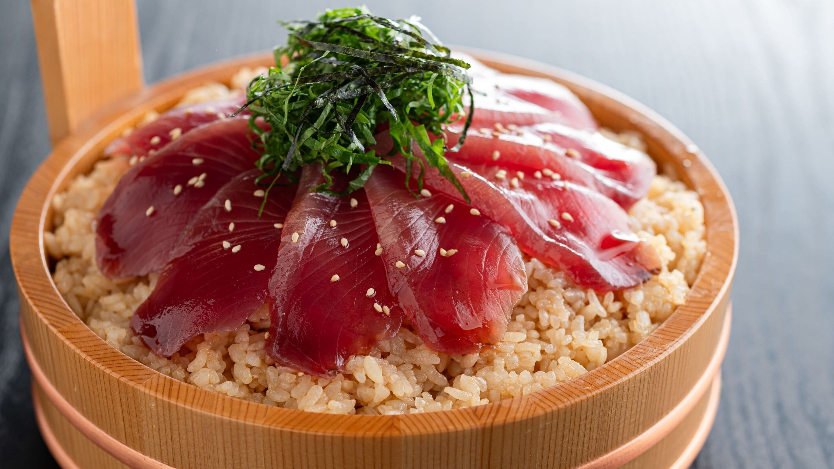 Uma tigela de madeira com arroz para sushi e fatias de peixe cru, cobertas com alga e sementes de gergelim.