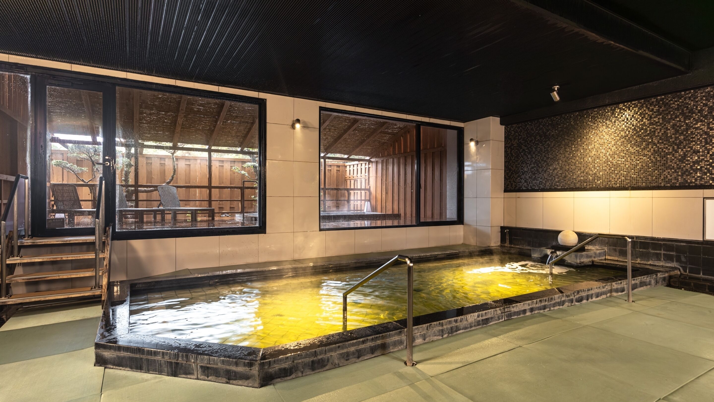 A hot spring bath area with a tiled floor, a small pool, and a wooden deck visible through the glass doors.