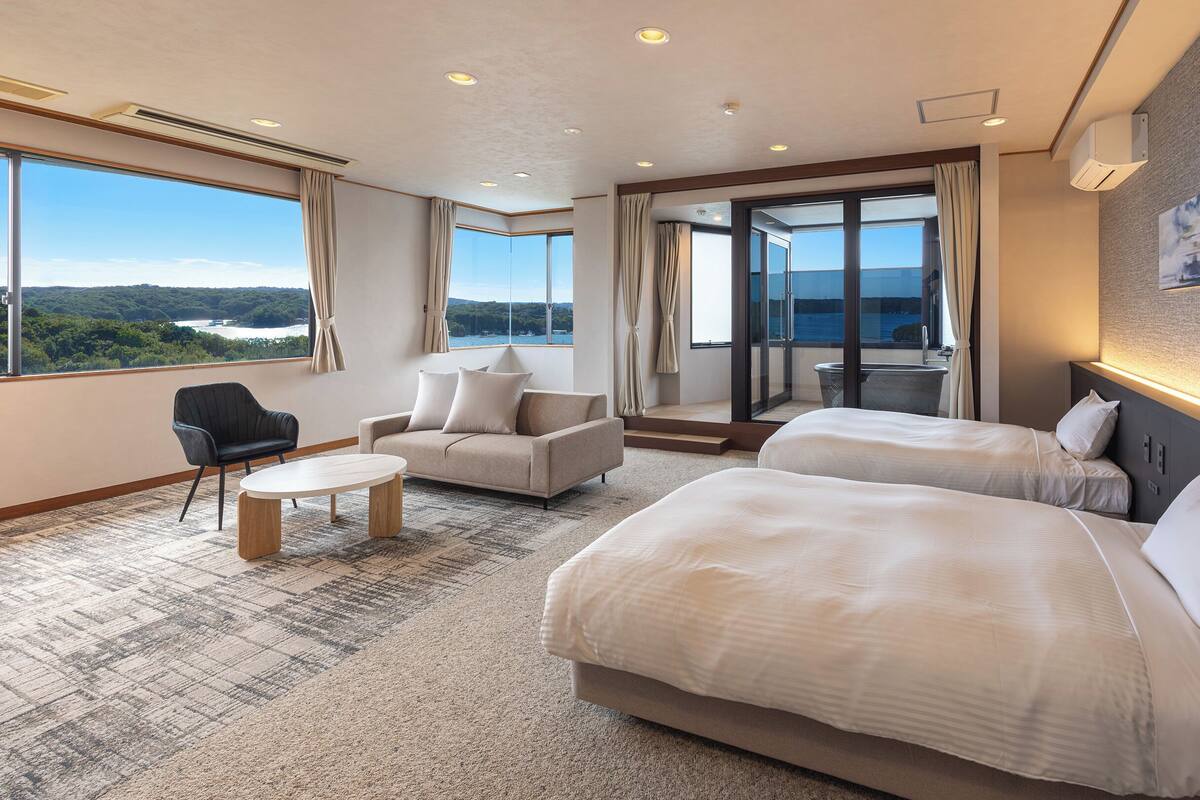 Ein Hotelzimmer mit zwei Betten, einem Sofa, einem kleinen Tisch und Blick ins Freie.