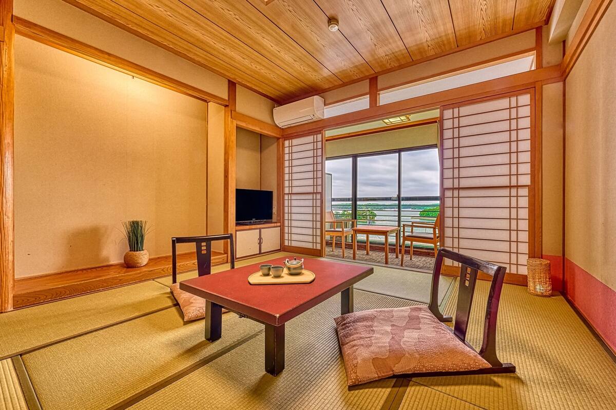 Ein traditioneller japanischer Raum mit Tatami-Boden, einem niedrigen Tisch mit Teeservice, einem Fernseher und Blick auf ein Gewässer.