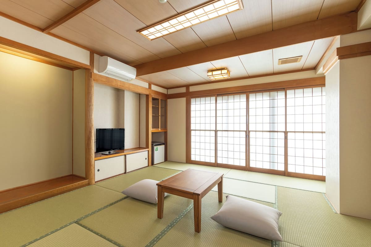 Ein traditioneller japanischer Raum mit Tatami-Bodenbelag, einem niedrigen Tisch, Kissen, einem Fernseher und Schiebetüren.