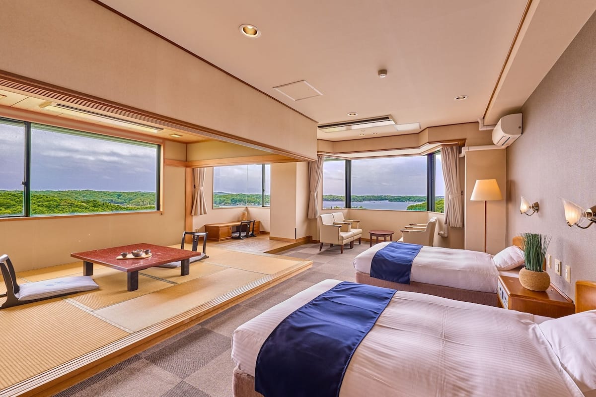 Ein Hotelzimmer mit zwei Betten, einem Esstisch, einem Schreibtisch und großen Fenstern mit Blick auf die Landschaft.