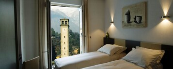 Quarto de hotel com duas camas, uma janela grande e vista para as montanhas, e uma torre de relógio visível lá fora.