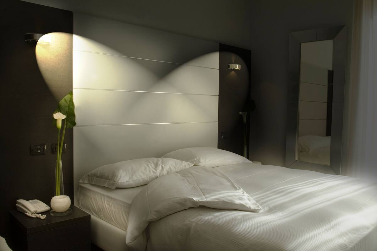 Un lit bien fait, avec du linge de lit blanc, une table de chevet avec un téléphone et un vase de fleurs, et un luminaire fixé au mur.