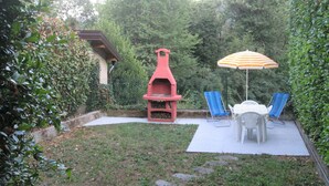 A backyard with a barbecue grill, two blue lawn chairs, a table with an umbrella, and a stone pathway. - La Casa del Borgo (Brissago-Valtravaglia)