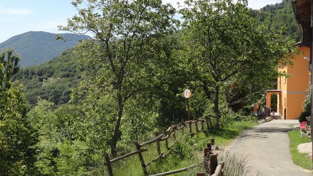 A rural road with a wooden fence, a building with a yellow facade, and a mountainous backdrop. - La Casa del Borgo (Brissago-Valtravaglia)