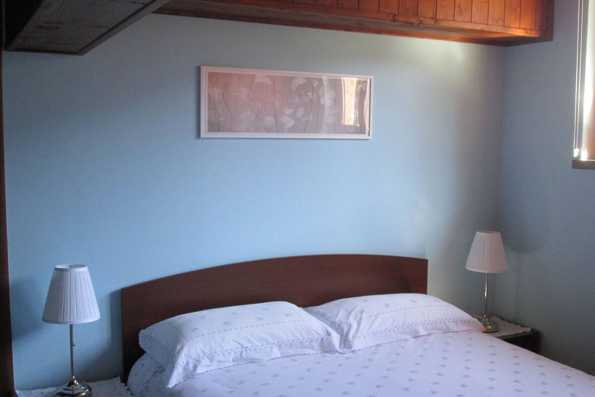 A bedroom with a wooden headboard, a bed with white linens, a nightstand with a lamp, and a wall-mounted picture.