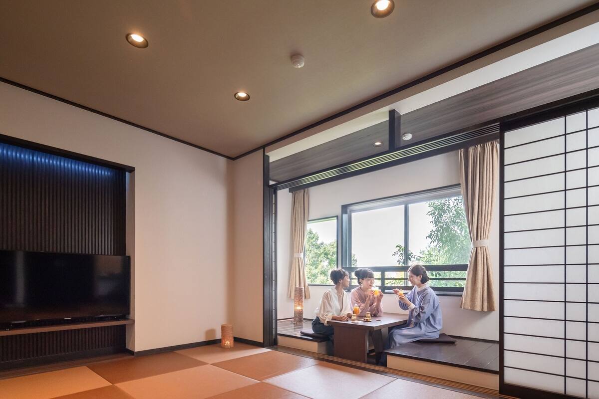 Una camera tradizionale giapponese con pavimento in tatami, una televisione, una porta scorrevole e vista sul verde circostante.