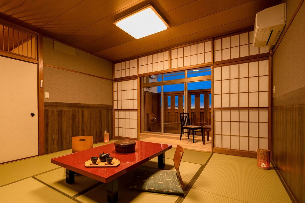 Una tradizionale stanza giapponese con pavimento in tatami, un tavolino con set per il tè, porte scorrevoli e vista attraverso una porta in vetro.