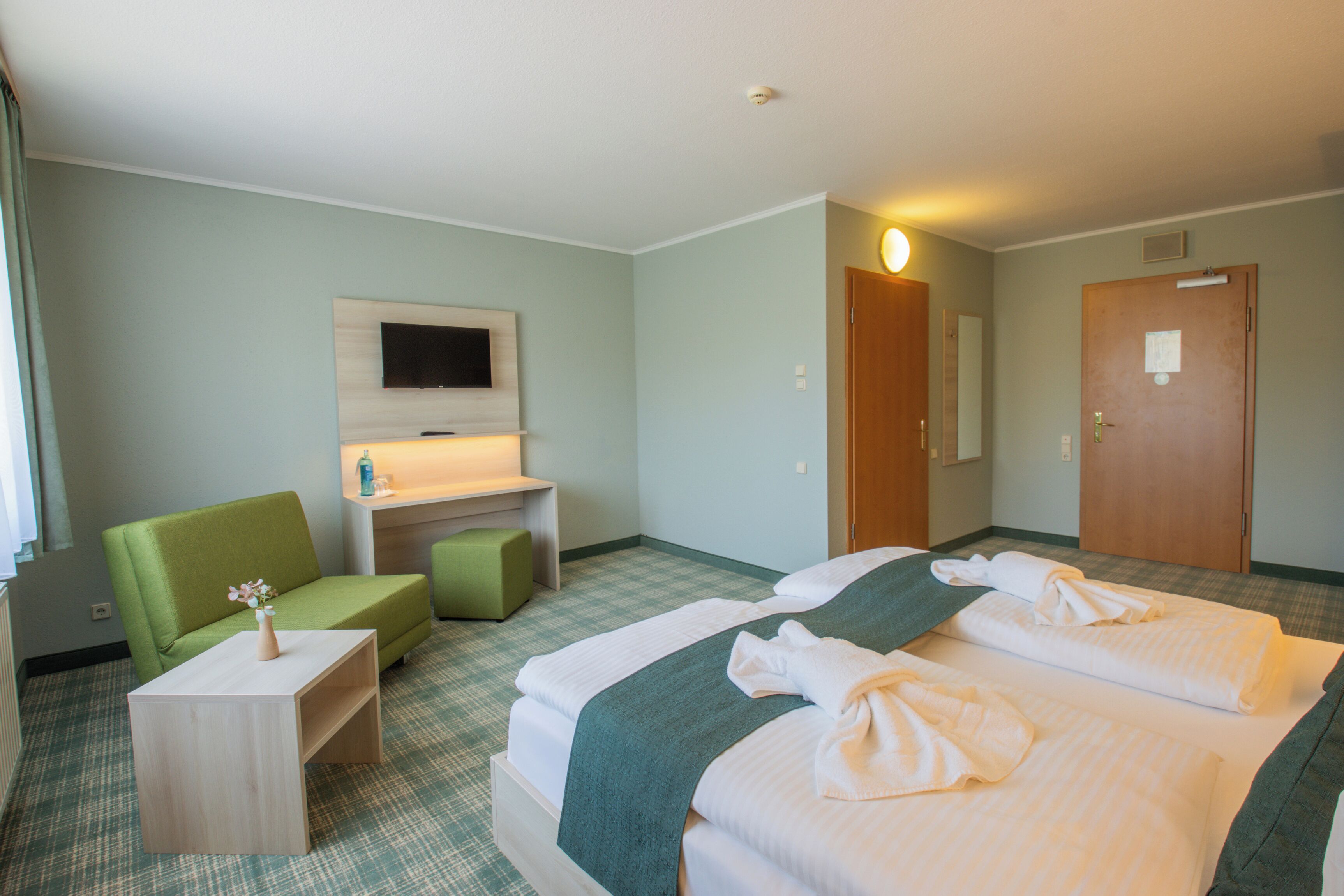 A hotel room with a bed, a green chair, a small table, a TV mounted on the wall, and a door.