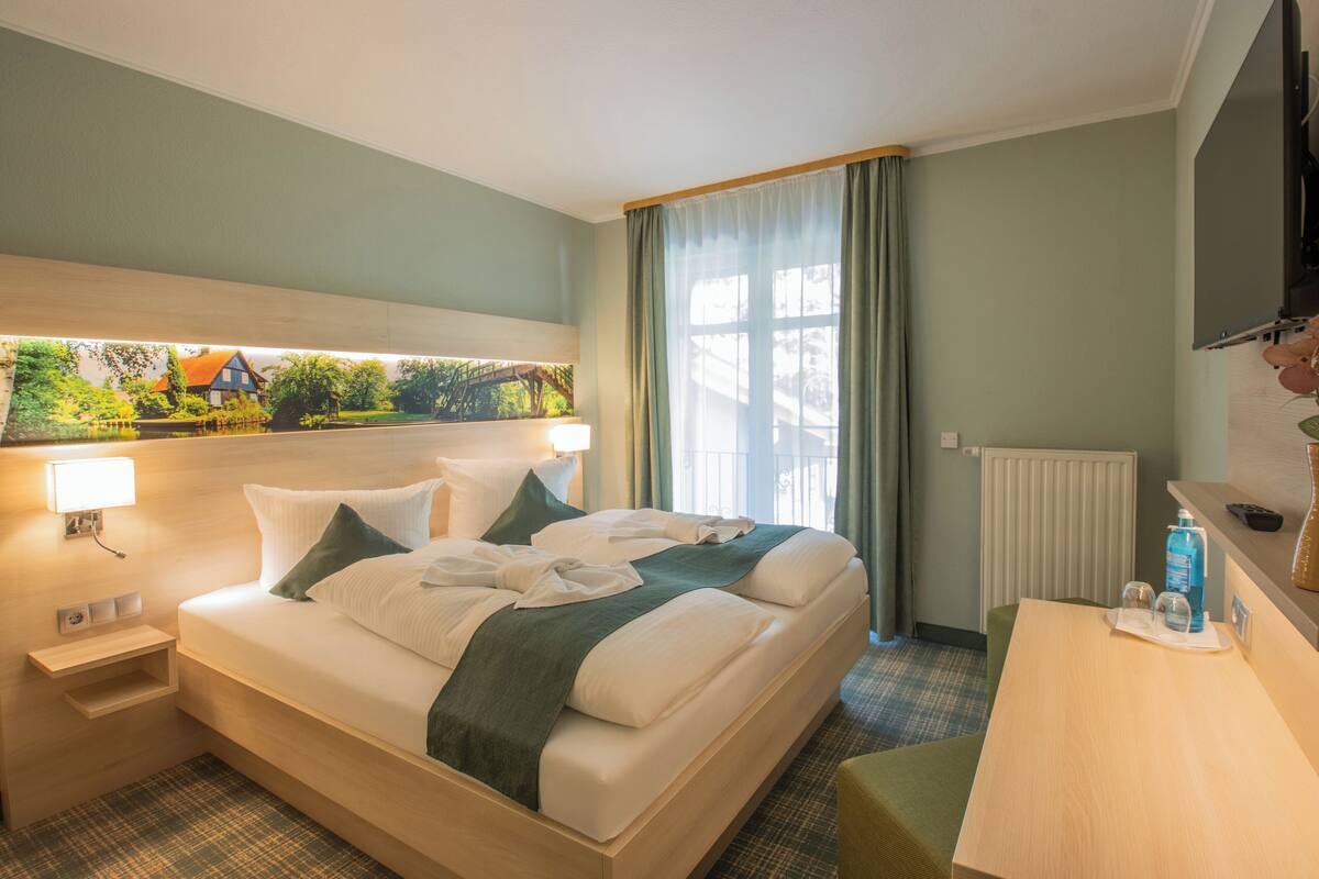 A hotel room with a bed, bedside table, lamp, and a view of greenery through the window.