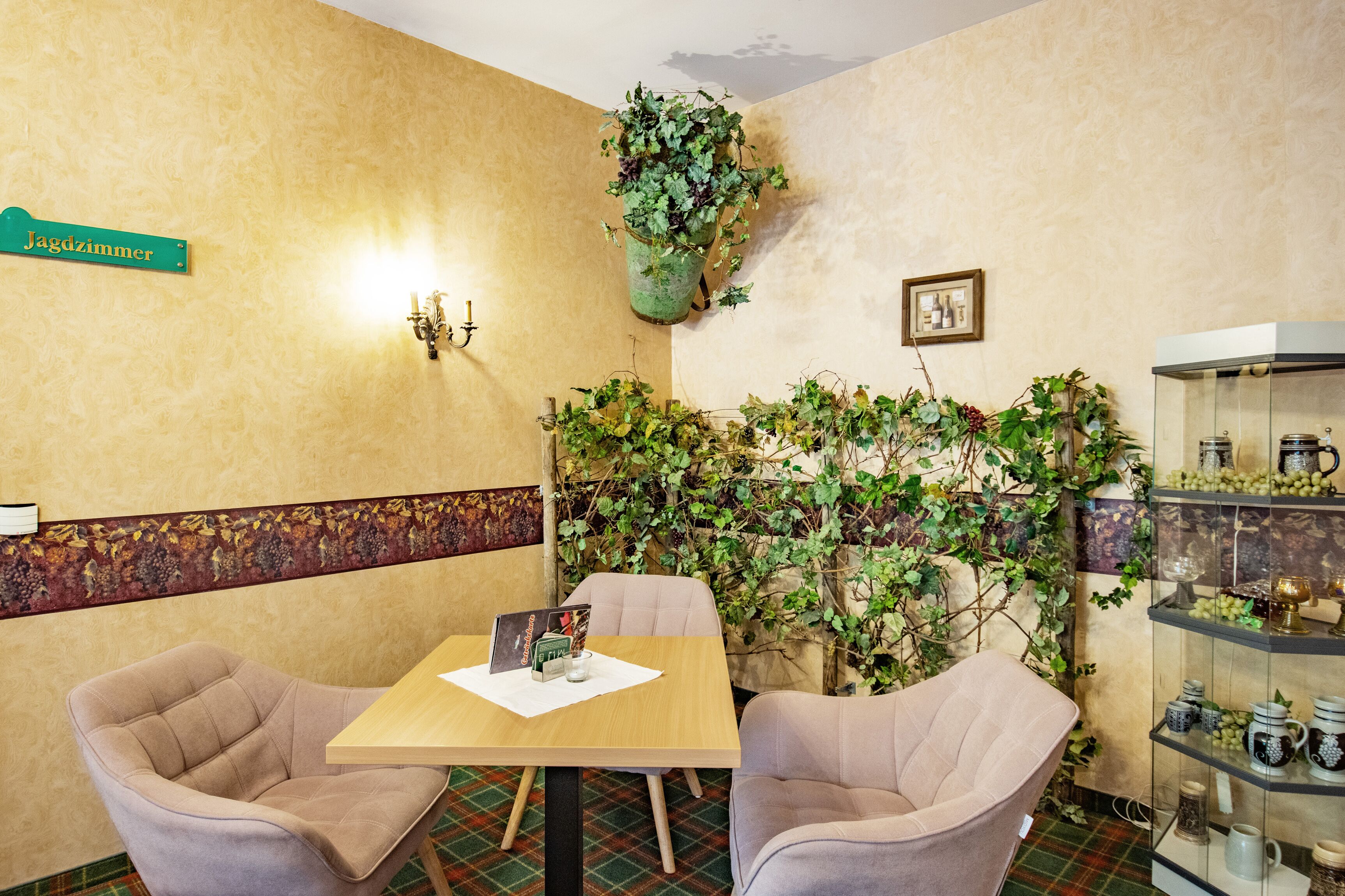 A room with a table and two chairs, a green sign, a wall-mounted light fixture, a potted plant, and a glass display case with items.
