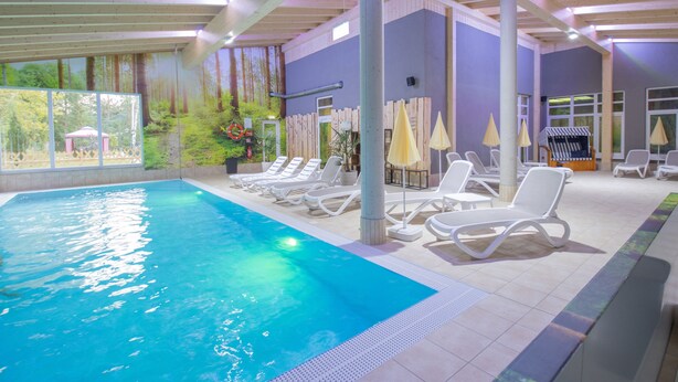 An indoor swimming pool with lounge chairs and umbrellas, surrounded by a wooden deck and large windows. - Ferien Hotel Spree-Neiße (Neuhausen/Spree)
