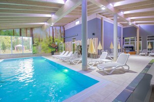 An indoor swimming pool with lounge chairs and umbrellas, surrounded by a wooden deck and large windows.