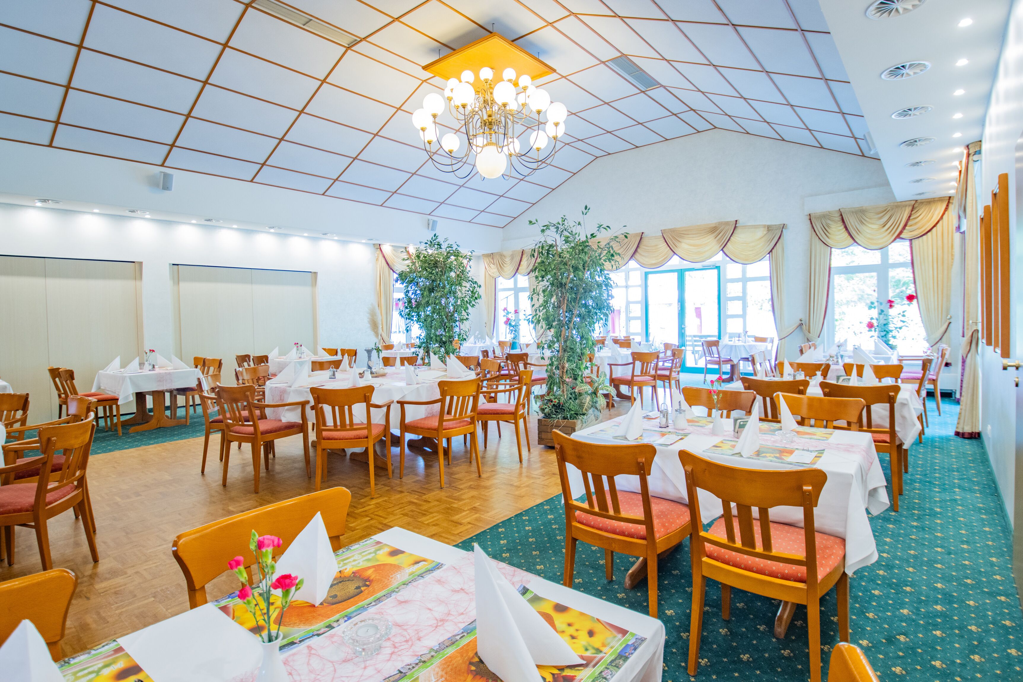 Ein geräumiger Essbereich mit mehreren Tischen und Stühlen, einer Chandelier und großen Fenstern mit Vorhängen.