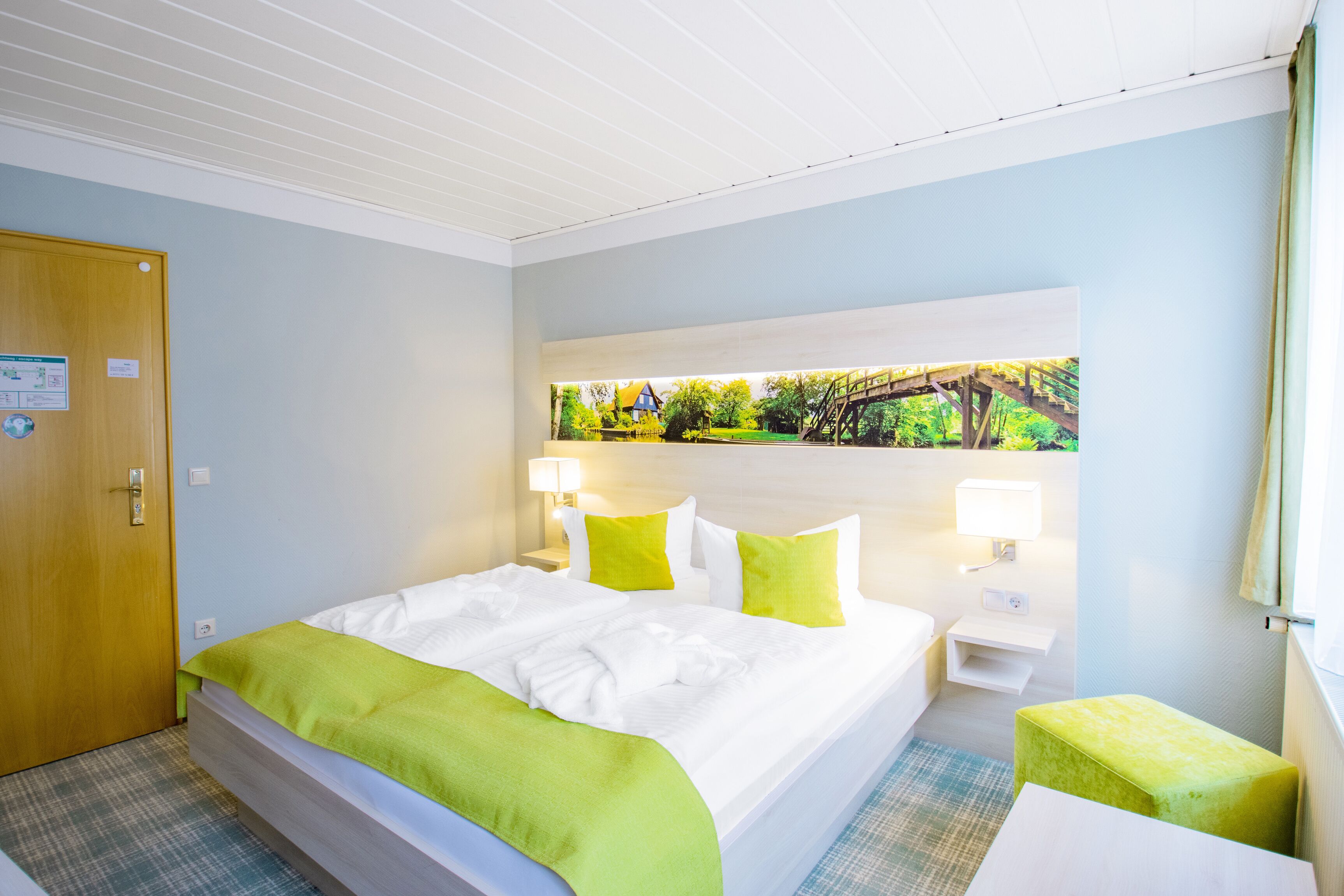 Habitación de hotel con cama, un banco verde, una mesita de noche, una lámpara y una ventana con vistas a zonas verdes.