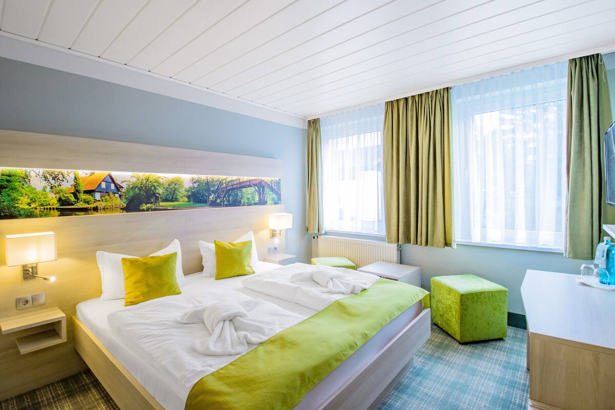 A hotel room with a bed, a desk, a chair, a window with curtains, and a painting on the wall.