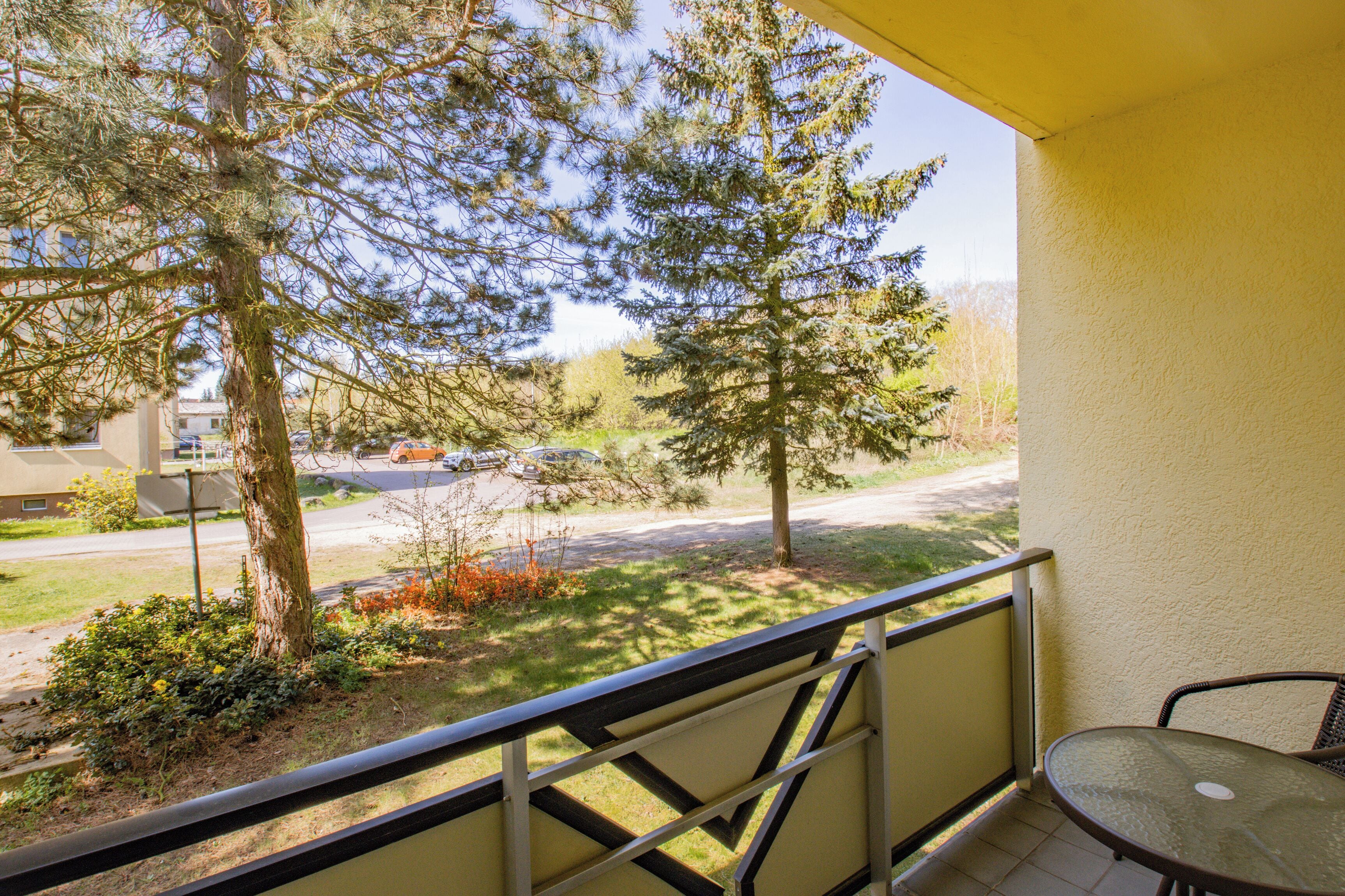 A balcony with a metal railing, a glass table, and a view of a road with cars and trees.